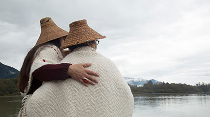 Two Tulalip Tribes members wearing traditional hats and blankets, embracing each other while standing near a serene lake with mountains in the background, representing cultural heritage and community